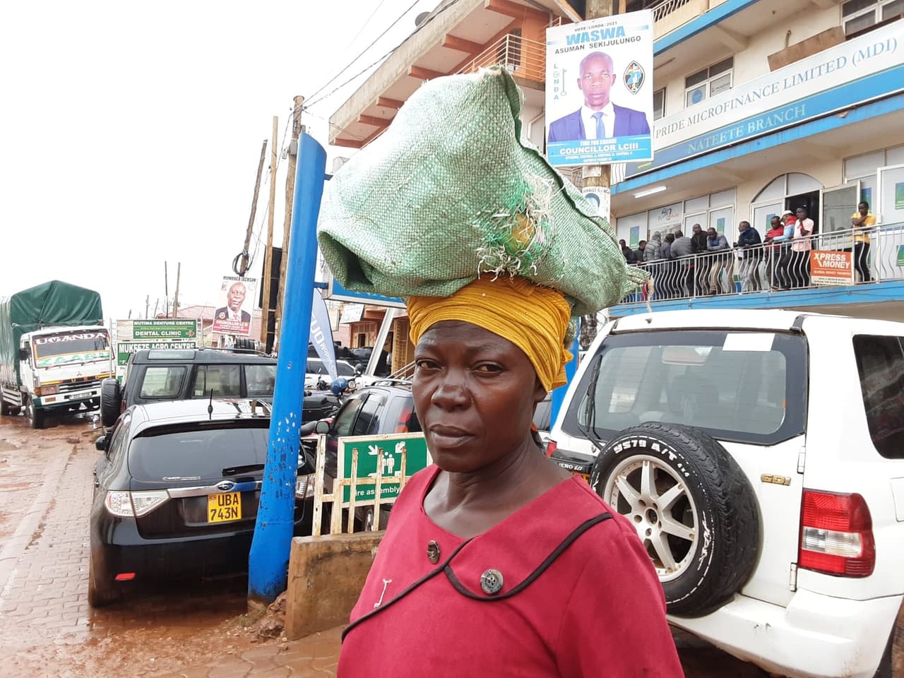 A trader balancing a full sack of produce on her head on a Nateete street, parked cars and a Pride Microfinance branch behind her.