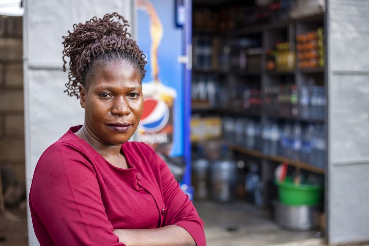 Florence standing with arms crossed outside her shop in Kampala, stocked shelves visible through the doorway behind her.