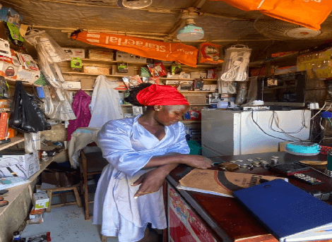 Nagujja Prossie standing at the counter of her mobile money kiosk in Kampala, surrounded by shelves of goods for sale.