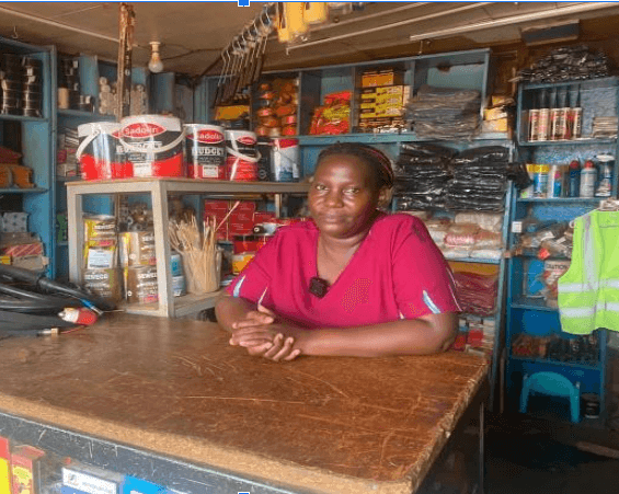 Namugerwa Sylvia at the counter of her hardware and household-goods shop in Kampala, shelves stocked behind her.